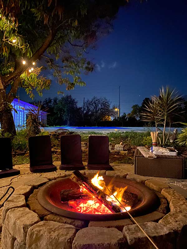 Evening photo of fire pit with flames in foreground, pool in background, under a starry sky.